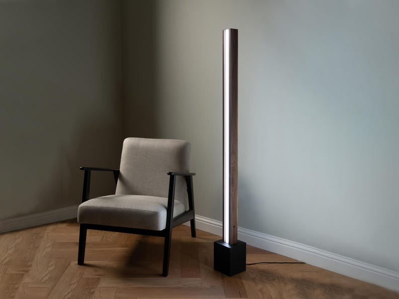 Pu&ograve; includere: Una lampada da terra moderna con una base in legno e una fonte di luce lunga e sottile. La lampada &egrave; accanto a una poltrona beige con gambe nere.