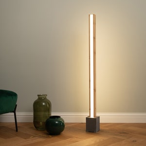 Może przedstawiać: Nowoczesna lampa stojąca z drewnianą ramą i betonową podstawą. Lampa ma długie, cienkie źródło światła, które emituje ciepłe białe światło.
