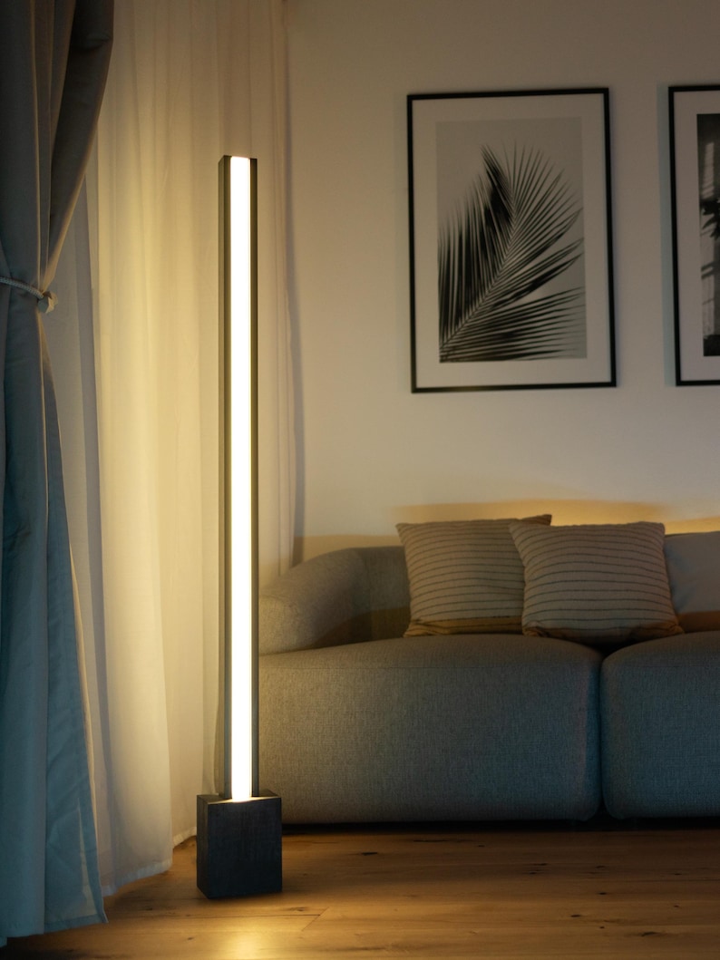 Pu&ograve; includere: Una lampada da terra moderna con un'alta struttura rettangolare e una base nera. La lampada illumina un soggiorno con un divano grigio e due cuscini.
