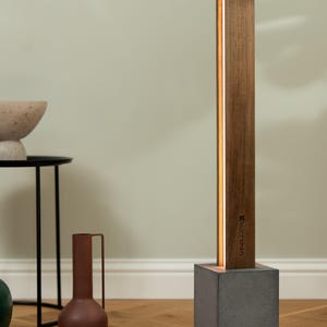 Pu&ograve; includere: Una lampada da terra moderna con una base in legno e una base in cemento. La lampada ha una sottile striscia di luce bianca che corre lungo il lato della base in legno. La lampada &egrave; etichettata "Norrsken".