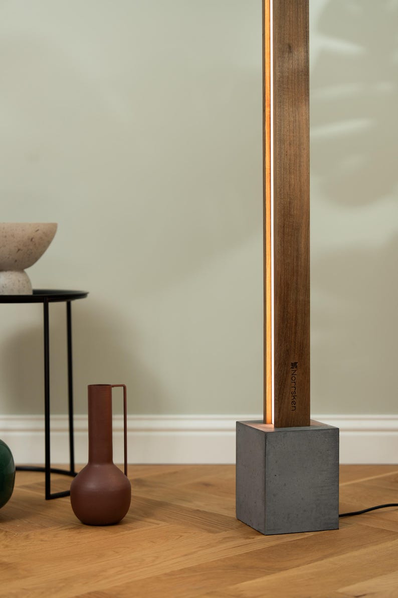 Pu&ograve; includere: Una lampada da terra moderna con una base in legno e una base in cemento. La lampada ha una sottile striscia di luce bianca che corre lungo il lato della base in legno. La lampada &egrave; etichettata "Norrsken".