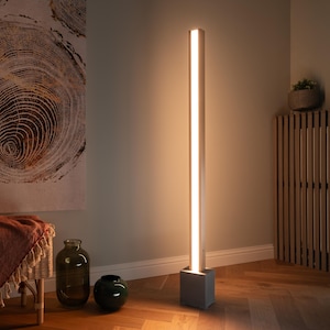 Pu&ograve; includere: Una lampada da terra moderna con una fonte di luce lunga, sottile e bianca e una base quadrata grigia. La lampada &egrave; accesa e proietta un bagliore caldo nell'area circostante.