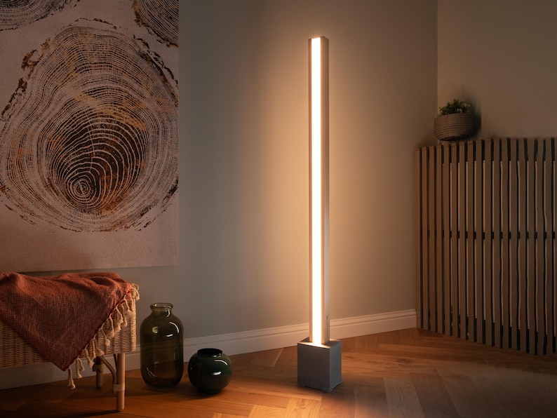 Pu&ograve; includere: Una lampada da terra moderna con una fonte di luce lunga, sottile e bianca e una base quadrata grigia. La lampada &egrave; accesa e proietta un bagliore caldo nell'area circostante.