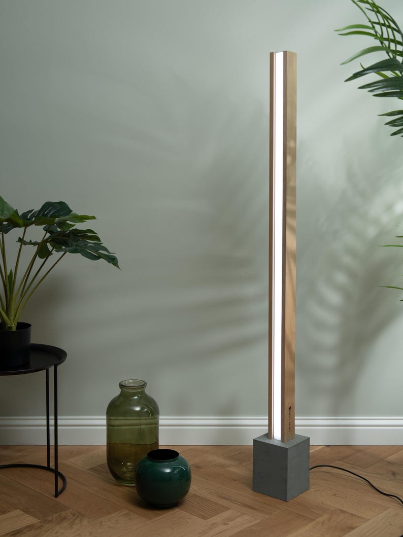 Pu&ograve; includere: Una lampada da terra moderna con una struttura in legno e una base in cemento. La lampada ha una fonte di luce bianca che illumina la stanza.