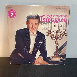 以下が含まれることがあります： リベラーチェをフィーチャーしたヴィンテージビニールレコードアルバムのカバー。カバーには、白い蝶ネクタイを着た黒いスーツ姿のリベラーチェの肖像画があります。アルバムのタイトルは「The Excitement of Mr. Showman Liberace」です。アルバムカバーはピンクとゴールドの配色です。