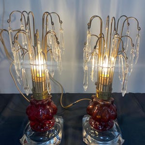 Puede incluir: Un par de lámparas de mesa antiguas con bases de vidrio rojo y acentos de cristal en forma de lágrima. Las lámparas tienen un marco de metal dorado y están cableadas para electricidad.