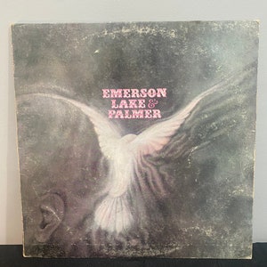 May include: A vintage vinyl record album cover featuring a white dove with outstretched wings against a dark gray background. The album title "Emerson Lake & Palmer" is printed in pink letters.