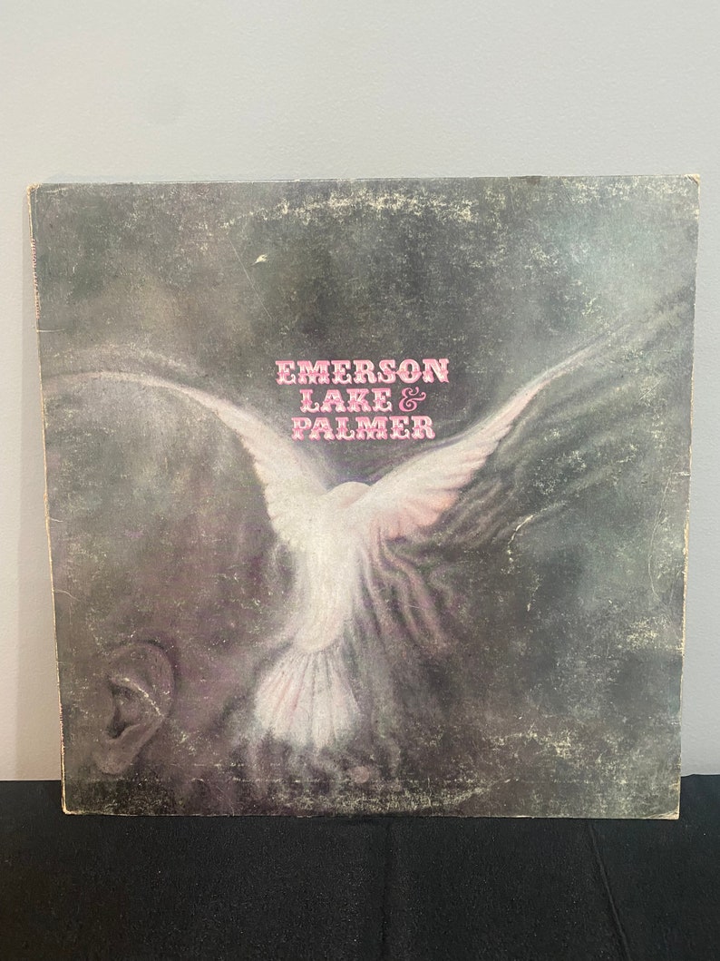 May include: A vintage vinyl record album cover featuring a white dove with outstretched wings against a dark gray background. The album title "Emerson Lake & Palmer" is printed in pink letters.