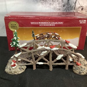 May include: A decorative Christmas bridge with a rustic wooden design, accented with faux snow and red bird figurines. The bridge is part of the "Santa's Workbench Collection" and is set against a winter scene backdrop.