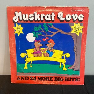 May include: A red, white, and blue record album cover with the title "Muskrat Love" in large, colorful letters. The cover features a cartoon illustration of two muskrats sitting on a yellow bench under a tree. The text "As seen on T.V." is in a star shape in the upper left corner. The text "2 Giant Records Only $3.99" is in a star shape in the upper right corner. The text "And 24 More Big Hits!" is at the bottom of the cover.