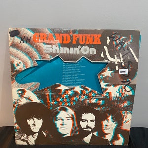 May include: A vintage vinyl record album cover featuring a band called "Grand Funk" with the album title "Shinin' On". The cover art features a blue and white star design with a 3D effect. The band members are pictured in the bottom half of the cover.
