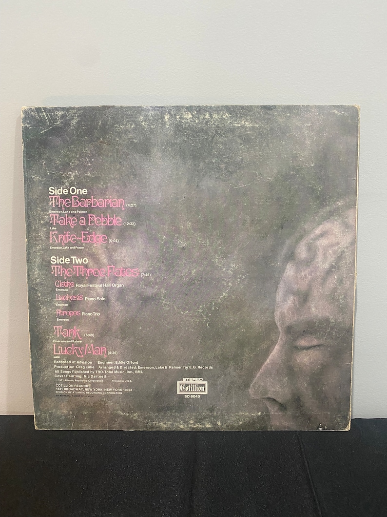 May include: Black and white album cover with a close-up of a person's face. The album is titled "Emerson, Lake & Palmer" and features the song titles "The Barbarian", "Take a Pebble", "Knife-Edge", "The Three Fates", "Clethe", "Lachesis", "Auropos", "Tank", and "Lucky Man".