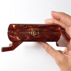 Può includere: Una piccola scatola di legno marrone con una chiusura dorata. La scatola ha un motivo decorativo a marmo e poggia su due piccole gambe.