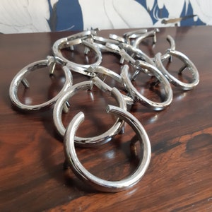 Può includere: Una collezione di pomelli ad anello in metallo argentato lucido, disposti su una superficie di legno scuro. Ogni pomello ha un design curvo, semicircolare, con un piccolo perno per il fissaggio. I pomelli sono probabilmente per cassetti o armadi.