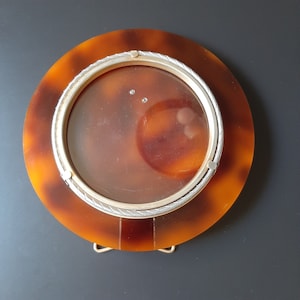 Könnte beinhalten: Ein runder Bilderrahmen aus Schildpatt mit einem silberfarbenen Rand. Der Rahmen hat einen klaren Glaseinsatz und ein dekoratives seilartiges Design am Rand.