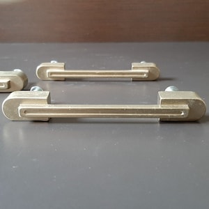 Könnte beinhalten: Drei goldfarbene Schrankgriffe mit rechteckiger Form und einer vertieften Mittellinie. Die Griffe haben ein modernes, minimalistisches Design.