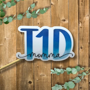 May include: A blue and white cake topper with the number "1" and the word "momma" in black script. The topper is on a wooden background.