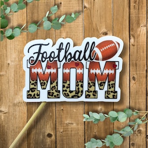 May include: White sign with the text "Football Mom" in black with a brown and white leopard print background. The sign has two footballs with red hearts on each side of the word "Mom".