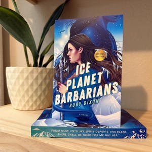 May include: A special edition of "Ice Planet Barbarians" by Ruby Dixon. The book cover features a woman with long brown hair holding a spear, set against a blue and white icy landscape. A quote is visible on the bottom of the book.