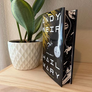 May include: A hardcover book titled "Project Hail Mary" by Andy Weir. The book has a black cover with white and yellow text and illustrations of astronauts and planets. The book is standing upright on a wooden surface next to a white geometric patterned pot with a green plant.