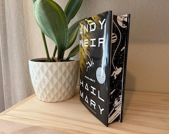 Op de afbeelding: Een hardcover boek getiteld "Project Hail Mary" van Andy Weir. Het boek heeft een zwarte kaft met witte en gele tekst en illustraties van astronauten en planeten. Het boek staat rechtop op een houten oppervlak naast een witte geometrisch gevormde pot met een groene plant.