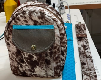 May include: A brown and white cowhide backpack with a turquoise trim and a brown leather flap. The backpack has a metal buckle and a small pocket on the front. The backpack is shown with two straps, one turquoise and one brown and white cowhide.