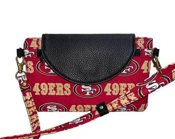 May include: A red and gold fabric crossbody bag with a black flap and a zipper closure. The bag has a strap with the words "49ers" printed on it.