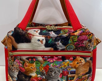 May include: A red and floral patterned handbag featuring a variety of kittens. The bag has red handles and trim, with a gold zipper and hardware. The main pattern includes kittens, flowers, and books.