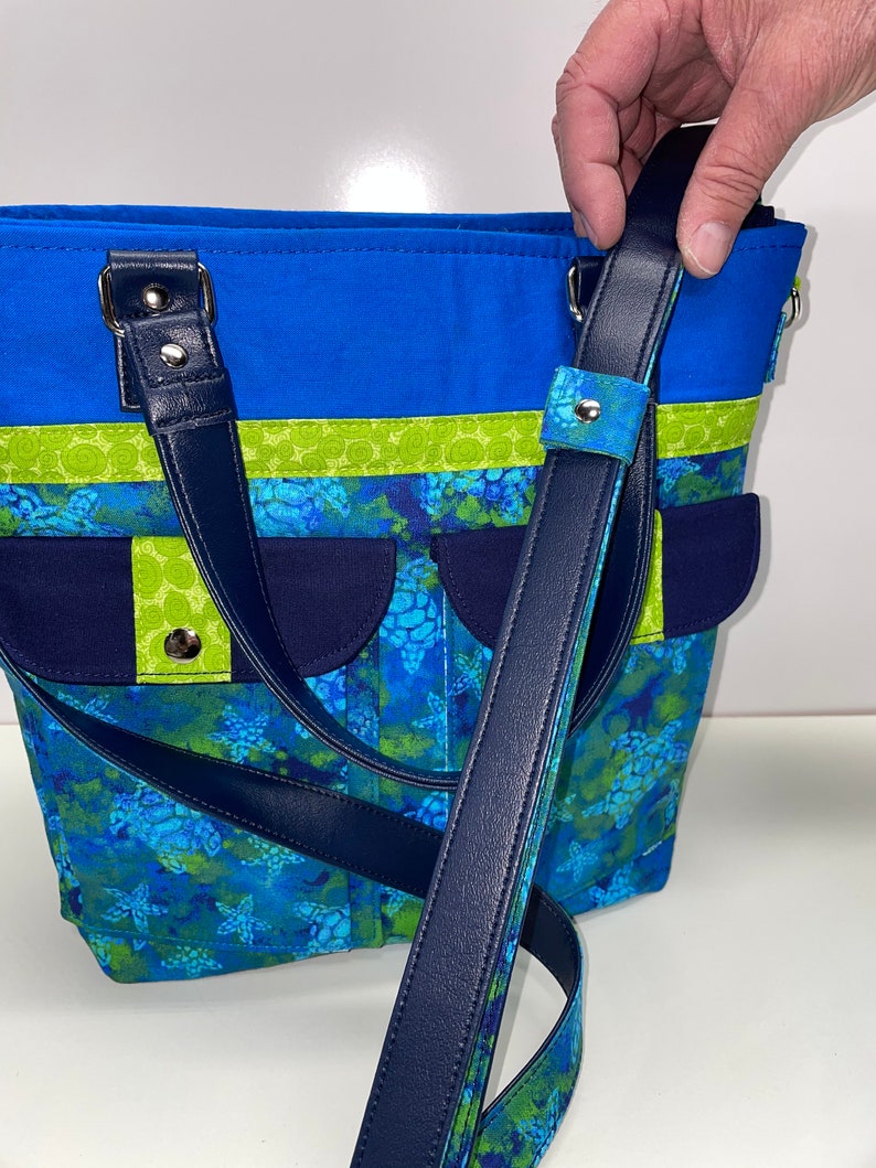 May include: A blue and green tote bag with a navy blue strap and a sea turtle print. The bag has a top zipper closure and two top handles.