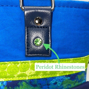 May include: Close-up of a blue fabric bag with a navy blue leather strap and a green peridot rhinestone. The bag has a green and blue patterned fabric lining.