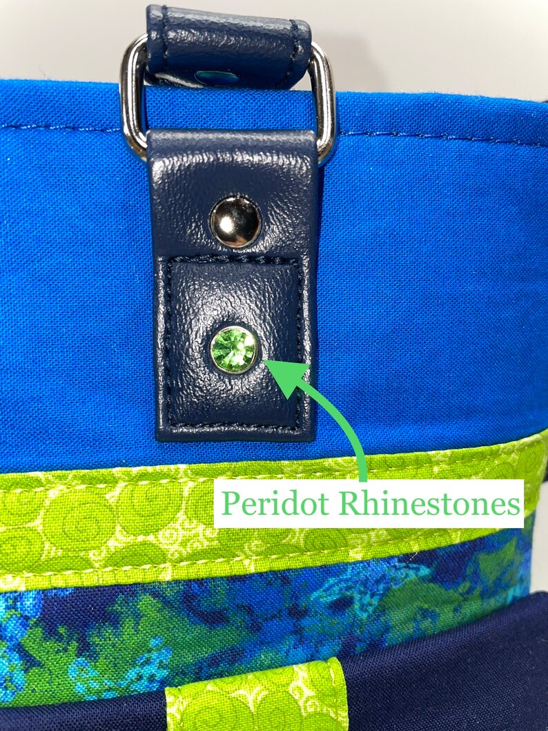 May include: Close-up of a blue fabric bag with a navy blue leather strap and a green peridot rhinestone. The bag has a green and blue patterned fabric lining.