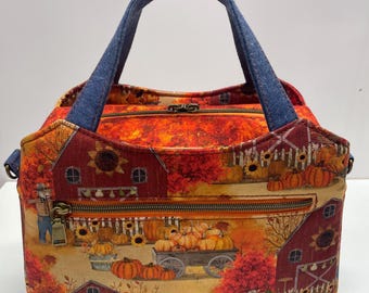 May include: A rectangular fabric handbag featuring a vibrant autumn harvest print with red barns, orange pumpkins, sunflowers, and scarecrows. The bag has a top zipper closure, a front zippered pocket, and two dark blue denim fabric handles.