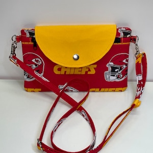 May include: Red and yellow fabric crossbody bag with a flap closure and a snap button. The bag has a white and yellow print of the Kansas City Chiefs logo and the word "CHIEFS".