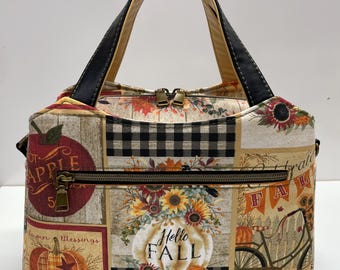 May include: A handbag featuring a patchwork design with autumn-themed prints including pumpkins, sunflowers, a bicycle, and apples. The bag has black handles, a top zipper closure, and a front horizontal zippered pocket with the text Hello Fall.