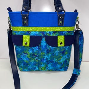 May include: A blue and green tote bag with a sea turtle print and a navy blue strap. The bag has a zipper closure and two pockets on the front.