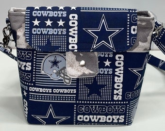 May include: A blue and white fabric crossbody bag with a patchwork design featuring the Dallas Cowboys logo, stars, and the words "Cowboys" repeated throughout.