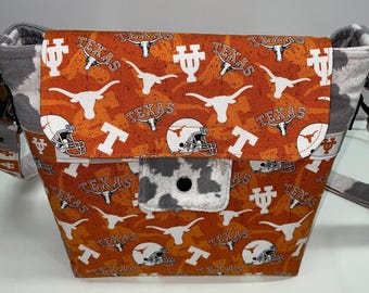 May include: A brown and orange fabric handbag with a white cow print pocket. The bag has a repeating pattern of the University of Texas Longhorns logo, a longhorn steer head, and the word "Texas".