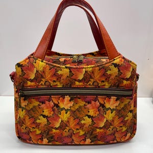 May include: A handbag featuring a vibrant autumn leaf pattern in shades of red, orange, and yellow. The bag has two sturdy reddish-brown handles, a top zipper closure with two pulls, and a horizontal front pocket with a dark brown zipper.