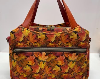 May include: A handbag featuring a vibrant autumn leaf pattern in shades of red, orange, and yellow. The bag has two sturdy reddish-brown handles, a top zipper closure with two pulls, and a horizontal front pocket with a dark brown zipper.