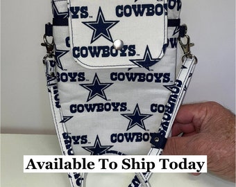 May include: White fabric phone crossbody bag with a blue and white Dallas Cowboys logo pattern. The bag has a zippered pocket and a long adjustable strap.