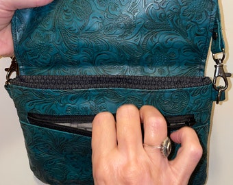 May include: Teal leather crossbody bag with a floral embossed design. The bag features a flap closure with a magnetic snap, a zippered pocket, and a shoulder strap. The interior lining is a patterned fabric.