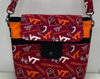 May include: A maroon and orange fabric tote bag with a black strap. The bag features a repeated pattern of the Virginia Tech logo, the words "Virginia Tech", and the word "Hokies".