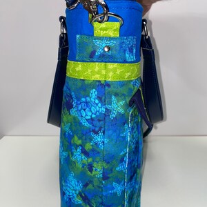May include: A blue and green tote bag with a sea turtle pattern. The bag has a navy blue strap and a green and blue pocket on the front.