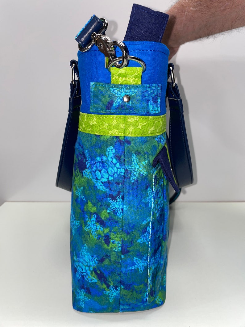 May include: A blue and green tote bag with a sea turtle pattern. The bag has a navy blue strap and a green and blue pocket on the front.