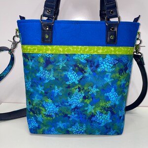 May include: A blue tote bag with a green and blue sea turtle print. The bag has a navy blue strap and a green and yellow trim.
