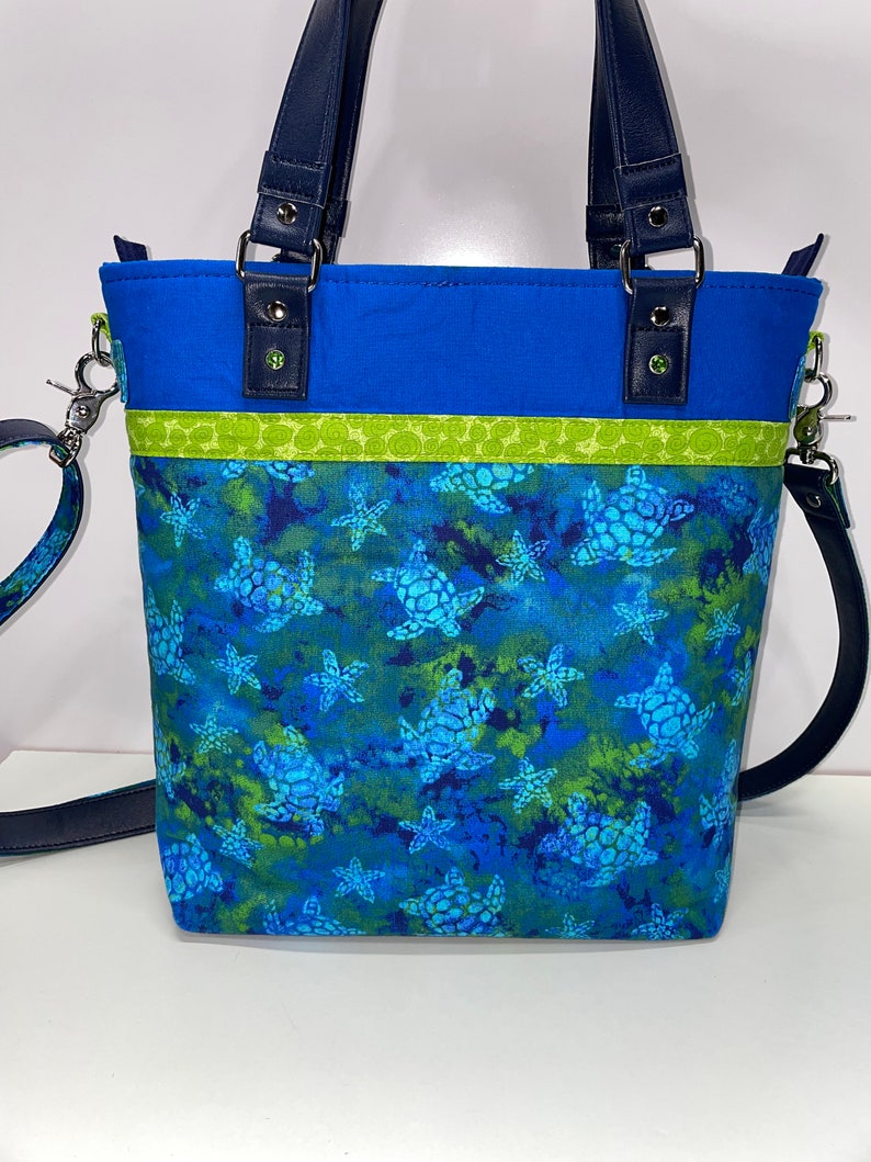 May include: A blue tote bag with a green and blue sea turtle print. The bag has a navy blue strap and a green and yellow trim.