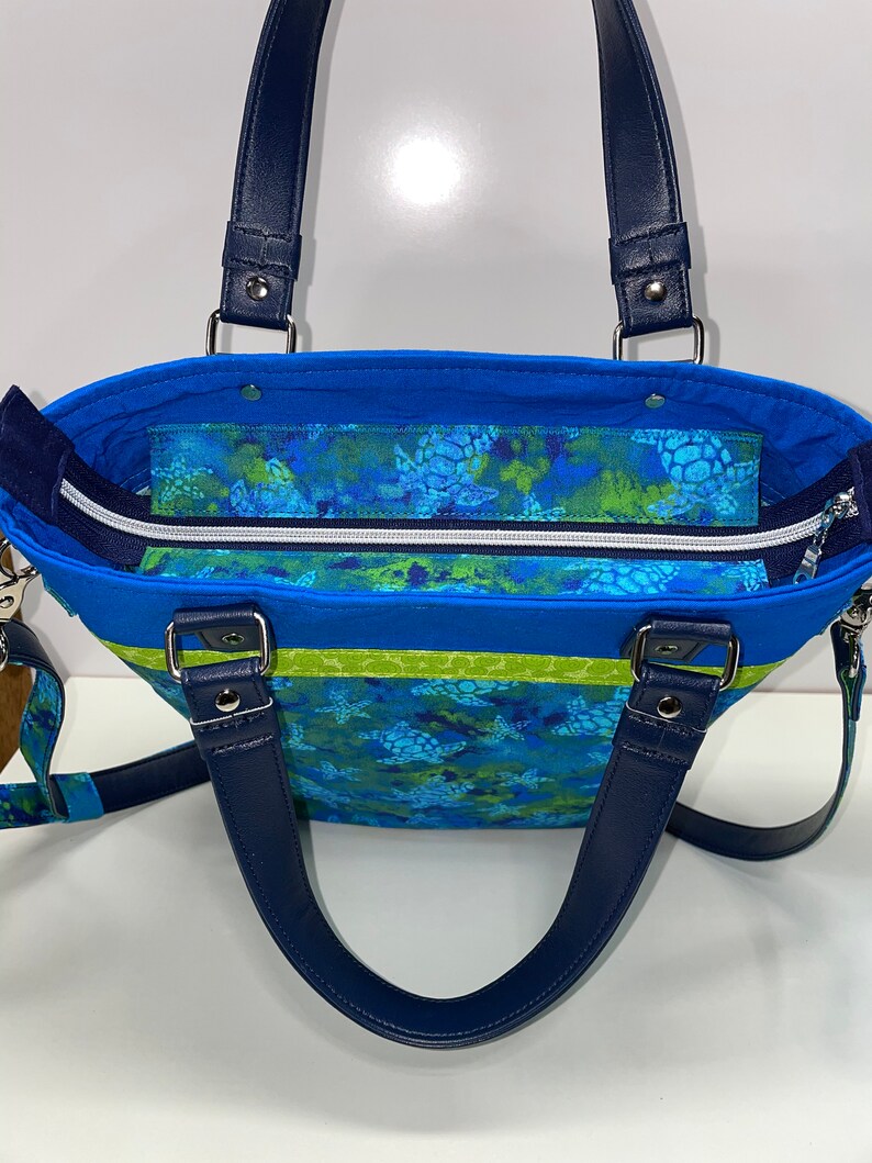 May include: A blue and green tote bag with a sea turtle print and navy blue handles. The bag has a zippered top closure and a lime green stripe.