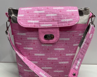 May include: Pink crossbody bag with a flap closure and a silver clasp. The bag is made of fabric with a white "Cowboys" print and stars. The strap is also pink with a white "Cowboys" print.