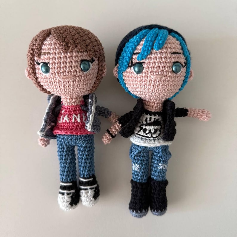 May include: Two handmade crochet dolls. One doll has brown hair, a red shirt, blue jeans, and a gray jacket. The other doll has blue-streaked black hair, a white shirt, blue jeans, and a black jacket. Both dolls have black and white shoes.
