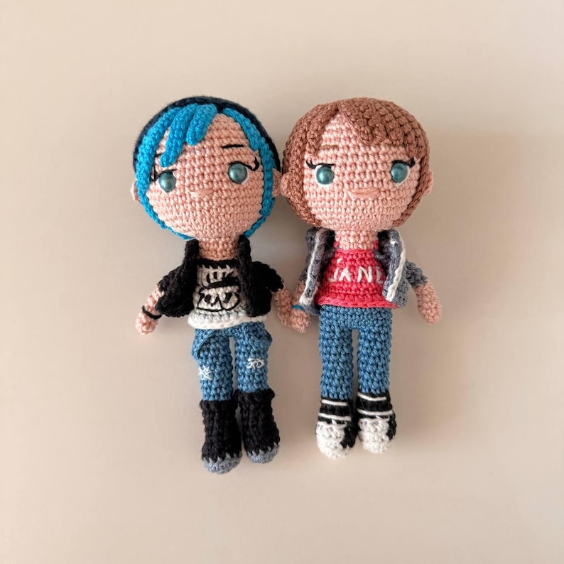 May include: Two handmade crochet dolls. One doll has blue hair, a black jacket, and blue jeans. The other doll has brown hair, a gray jacket, a red shirt with "JAN" on it, and blue jeans. Both dolls have black boots.